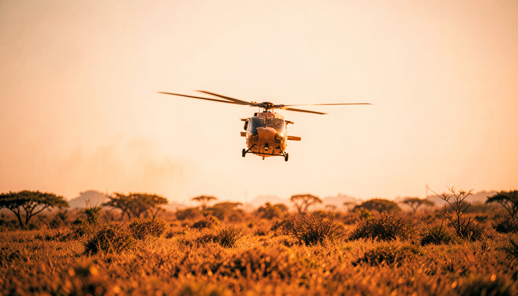 A helicopter flying low over savannah terrain in hazy midday light, wide cinematic frame