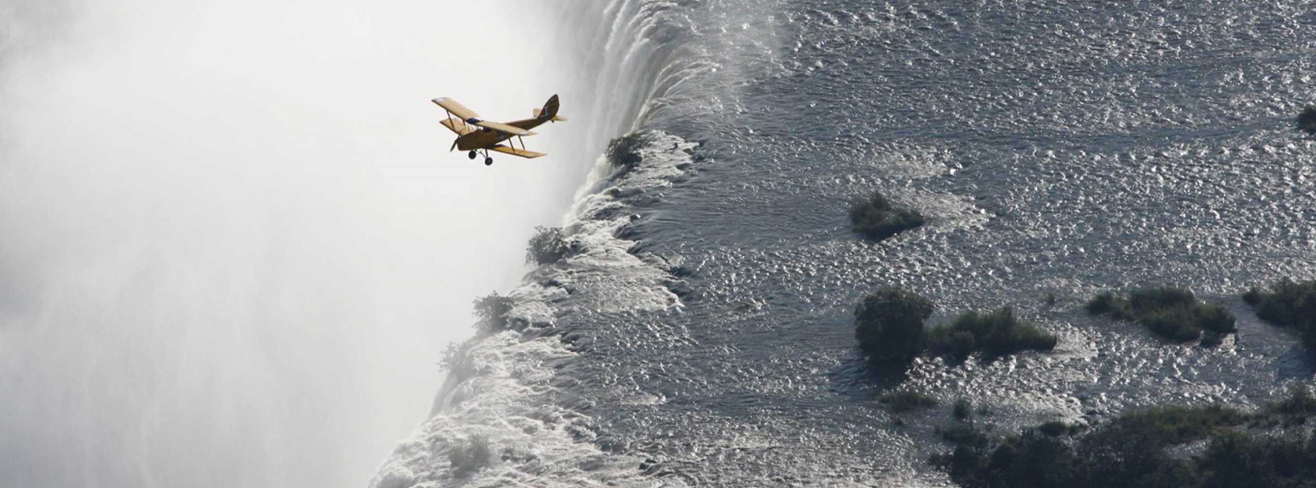 Small biplane flying over the edge of a large waterfall with mist rising, grey and white tones, dramatic aerial scene