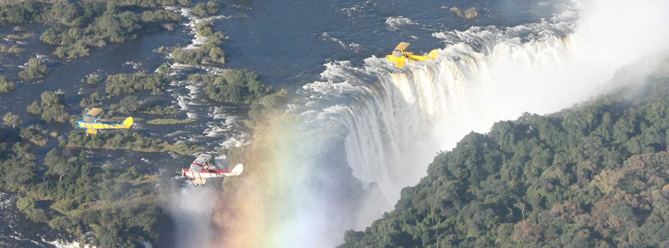 Three biplanes in yellow and blue flying in formation over a wide waterfall with rainbow in the mist, lush green forest below